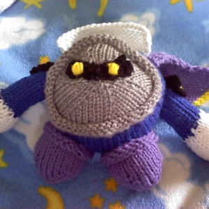 May include: A knitted plush toy resembling a character from a video game. The toy is grey, purple, and white with yellow eyes.