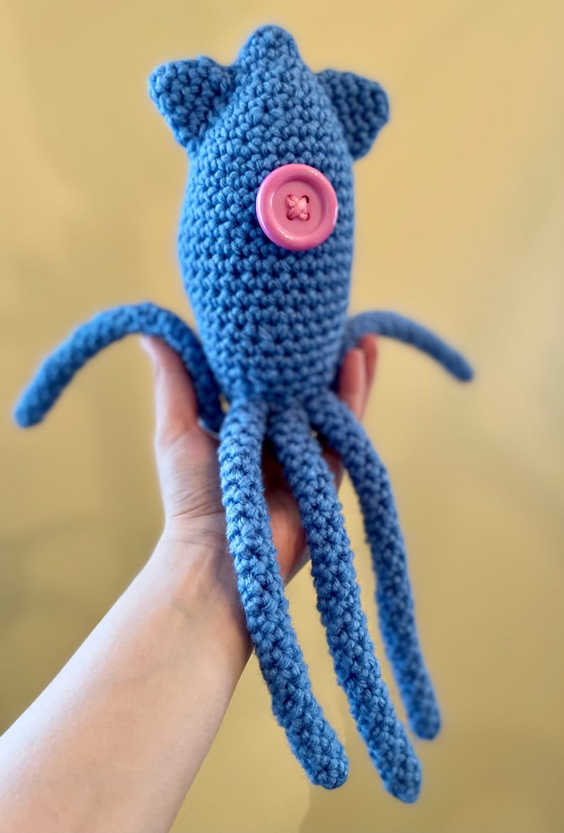 Pu&ograve; includere: Un giocattolo di calamaro all'uncinetto blu con un bottone rosa per un occhio. Il calamaro ha otto tentacoli.