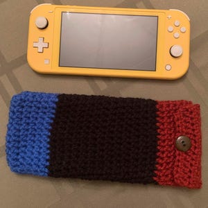 May include: A crocheted case for a Nintendo Switch console. The case is made of blue, black, and red yarn and has a button closure.