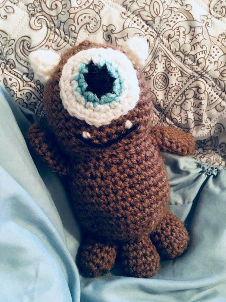 May include: A brown crocheted stuffed toy with a single white eye with a blue and black centre. The toy is lying on a blue blanket.
