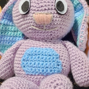 May include: A crocheted plush toy bunny with purple fur, blue and green ears, and black eyes. The bunny has a pink nose and a blue patch on its chest.