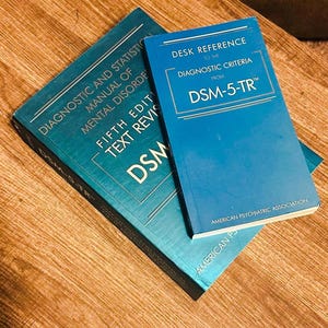 以下が含まれることがあります： 木製の表面に2冊の本。1冊はティール色で、「Diagnostic and Statistical Manual of Mental Disorders, Fifth Edition, Text Revision, DSM-5-TR」と題されています。もう1冊は青色で、「Desk Reference to the Diagnostic Criteria from DSM-5-TR」と題されています。