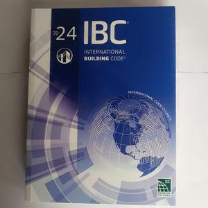 May include: A hardcover book titled "2024 IBC International Building Code." The cover is primarily blue with a graphic of a globe and the International Code Council logo. The book is a reference for building codes.