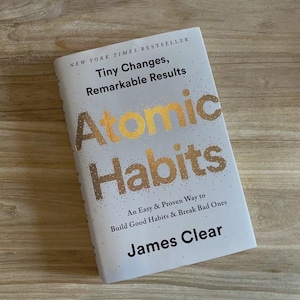 May include: A hardcover book titled "Atomic Habits" by James Clear. The book has a white cover with gold lettering and the text "Tiny Changes, Remarkable Results." The book is on a wooden surface.