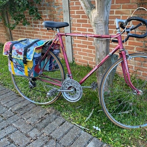 May include: A vintage pink bicycle with a black seat and handlebars. The bicycle has a colorful patterned pannier bag attached to the rear. The tires have a tan sidewall. The bike is parked next to a brick wall and a tree.