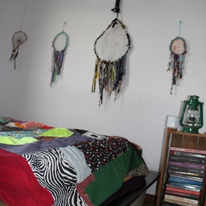 Może przedstawiać: Kołdra patchworkowa z wzorem zebry, zielonym, czerwonym i czarnym materiałem, na ramie ł&oacute;żka z białą głową. Na ścianie za ł&oacute;żkiem wiszą cztery łapacze sn&oacute;w z białymi, niebieskimi i zielonymi akcentami. Drewniana p&oacute;łka z książkami i latarnią znajduje się po prawej stronie obrazu.