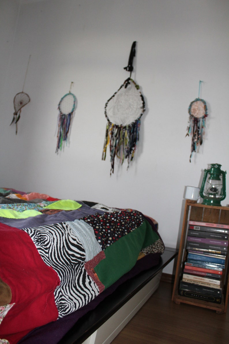 Może przedstawiać: Kołdra patchworkowa z wzorem zebry, zielonym, czerwonym i czarnym materiałem, na ramie ł&oacute;żka z białą głową. Na ścianie za ł&oacute;żkiem wiszą cztery łapacze sn&oacute;w z białymi, niebieskimi i zielonymi akcentami. Drewniana p&oacute;łka z książkami i latarnią znajduje się po prawej stronie obrazu.