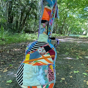 Könnte beinhalten: Ein farbenfroher Gitarrenkoffer mit Patchwork-Design. Der Koffer weist eine Vielzahl von Stoffen auf, darunter Jeansstoff, Blumenmuster und Streifen. Er hat einen Reißverschluss und eine lila Umrandung. Der Koffer steht auf einem Weg, der von Bäumen gesäumt ist.