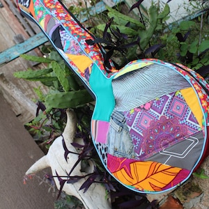 Puede incluir: Una funda de guitarra colorida hecha de tela de patchwork con una variedad de patrones, incluyendo denim, flores y diseños geométricos. La funda es azul, naranja, rosa y gris.