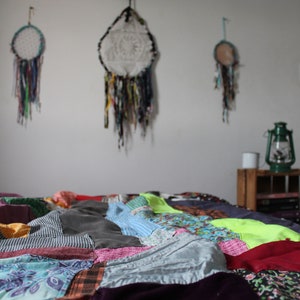 Może przedstawiać: Kolorowa kołdra patchworkowa z r&oacute;żnymi tkaninami i wzorami, w tym kwiatowymi, kratowymi i w paski. Kołdra leży na ł&oacute;żku w pokoju z białymi ścianami i łapaczami sn&oacute;w wiszącymi na ścianie.