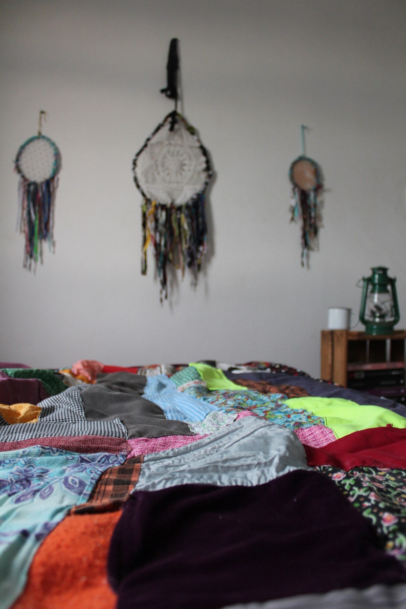 Może przedstawiać: Kolorowa kołdra patchworkowa z r&oacute;żnymi tkaninami i wzorami, w tym kwiatowymi, kratowymi i w paski. Kołdra leży na ł&oacute;żku w pokoju z białymi ścianami i łapaczami sn&oacute;w wiszącymi na ścianie.