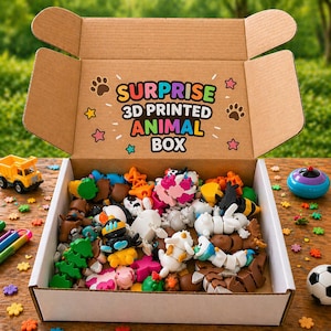 Puede incluir: Una caja de cartón abierta llena de varias figuras de animales impresas en 3D. La caja está etiquetada como "SURPRISE 3D PRINTED ANIMAL BOX" en texto colorido. Las figuras son de varios colores y formas, incluyendo una abeja, un perro y una vaca.