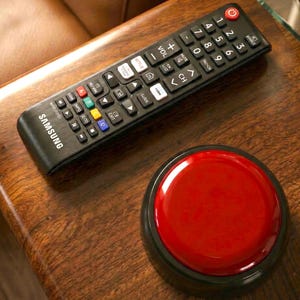 May include: A black Samsung remote control with numerous buttons and a large red button. The remote is on a wooden surface next to a large, round, red button with a black base. The remote has buttons for Netflix, volume, and channel selection.
