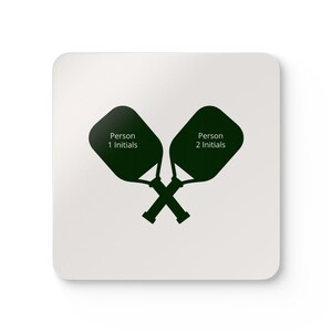 Könnte beinhalten: Ein quadratischer weißer Untersetzer mit einer dunkelgrünen Grafik von zwei gekreuzten Pickleball-Schlägern. Jeder Schläger hat eine dunkelgrüne ovale Form mit dem Text "Person 1 Initialen" und "Person 2 Initialen" in Weiß.