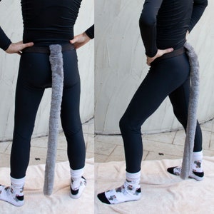 Könnte beinhalten: Eine Person trägt schwarze Leggings und Socken mit einem grauen, pelzigen Schwanz, der an einem schwarzen Gürtel befestigt ist.