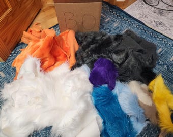 May include: A collection of assorted faux fur fabric scraps in various colors including white, orange, black, purple, bright blue, light blue, tan, and yellow. A brown cardboard box with the letters BO written in black marker sits behind the fabric.