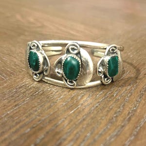 May include: A silver cuff bracelet featuring three oval, deep green gemstones. The bracelet has an open design with decorative swirls and leaf-like accents around each stone. The bracelet is displayed on a wooden surface.