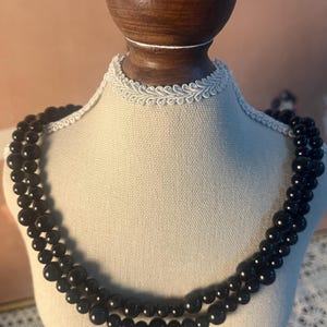 May include: A two-strand black beaded necklace displayed on a mannequin. The beads are round and glossy, with a consistent size throughout each strand. The mannequin has a cream-colored fabric and a white lace trim.
