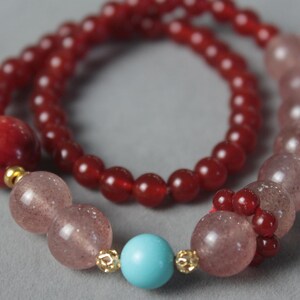 May include: A necklace featuring two strands of beads in shades of red and pink. The necklace includes large, round red beads, smaller red beads, and pinkish-red beads. A turquoise bead and gold-colored accents are also present.
