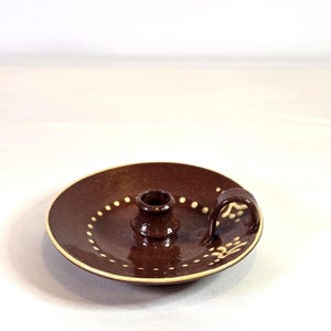Peut inclure: Un bougeoir en céramique marron fait à la main, avec une base circulaire et une petite poignée. Le bougeoir comporte un puits central pour la bougie et est décoré d'accents pointillés blancs autour du bord.