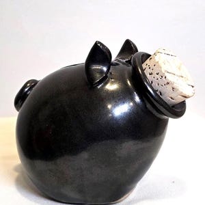 May include: A black ceramic piggy bank with a cork stopper. The piggy bank has a glossy finish and features small ears and a slot for inserting coins. The cork is light brown and fits snugly in the opening.