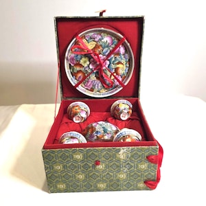 Puede incluir: Un juego de té decorativo en una caja. El juego incluye una bandeja, una tetera y varias tazas de té. La bandeja y las tazas presentan diseños florales con acentos rojos, rosas y amarillos. La caja es verde con un interior rojo.