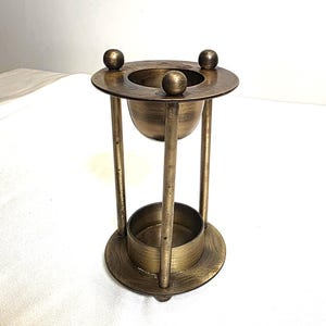Könnte beinhalten: Ein Vintage-Räuchergefäß aus Messing. Es hat einen kreisförmigen Sockel und eine kreisförmige Oberseite mit drei kleinen Kugelakzenten auf der Oberseite. Die zentrale Schale wird von drei vertikalen Stäben getragen, darunter ein zylindrischer Sockel.