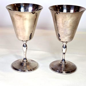 Puede incluir: Dos copas de plata con bordes acampanados y tallos delgados. Las copas tienen una superficie pulida y reflectante y se asientan sobre bases redondas. El tallo tiene un diseño bulboso decorativo. Las copas son probablemente para servir bebidas.