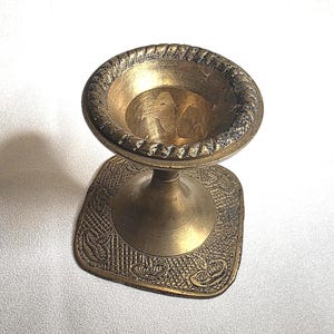 Könnte beinhalten: Ein Vintage-Kerzenhalter aus Messing mit quadratischem Sockel und rundem, strukturiertem Oberteil. Der Kerzenhalter hat einen dekorativen, seilartigen Rand um die Oberseite und komplizierte Muster auf dem Sockel. Das Messing hat eine warme, gealterte Patina.