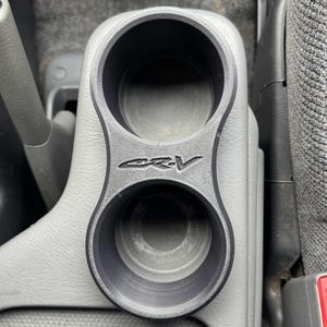 Op de afbeelding: Een zwarte dubbele bekerhouder met de letters "CRV" in reliëf op de voorkant. De bekerhouder is ontworpen om in de middenconsole van een auto te passen. De binnenkant van de bekerhouders is zwart en de buitenkant is lichtgrijs.