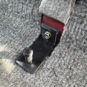 May include: Close-up of a black 3D-printed bracket with a silver bolt and a cylindrical post. The bracket is attached to a gray, textured fabric, with a section of red material visible beneath the bracket.