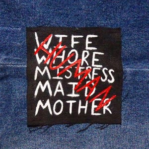 May include: A black square patch with white text reading "WIFE WHORE MISTRESS MAID MOTHER". The words "WHORE", "MISTRESS", and "MAID" are crossed out in red. The patch is sewn onto blue denim fabric.