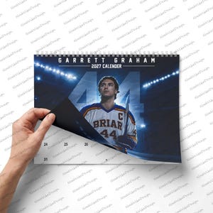 May include: A 2027 wall calendar featuring a hockey player in a white and blue Briar jersey with the number 44 and a captain's C. The text reads Garrett Graham 2027 Calender. The image shows a stadium background with bright overhead arena lights.