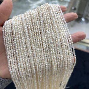 May include: A collection of creamy white, small, irregular-shaped pearls strung together. The pearls are held in a hand, showcasing their delicate texture and natural luster. The strands appear to be ready for jewelry making.