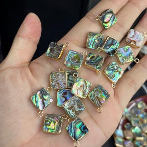 May include: A collection of square-shaped abalone shell charms, each featuring a gold-toned metal border and a small loop at the top. The iridescent shell surfaces display a vibrant mix of green, blue, purple, and pink hues with natural patterns.