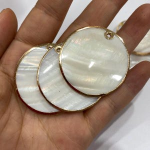 May include: Round, mother-of-pearl pendants with a gold-colored metal rim. Each pendant has a small hole at the top for stringing. The iridescent white surface of the pendants catches the light, creating a shimmering effect. These pendants are ideal for jewelry making.