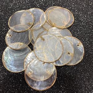 May include: A pile of iridescent, circular shell pendants with gold-coloured edging. Each pendant has a small hole for threading. The shells have a pearlescent sheen, reflecting light with a range of colours.