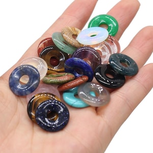 May include: A collection of assorted gemstone donut pendants in various colors, including green, blue, brown, and clear. The pendants are round with a hole in the center, and are held in a hand.