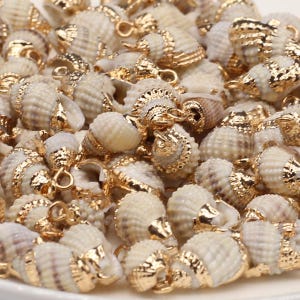Könnte beinhalten: Eine Ansammlung kleiner, natürlicher cremeweißer Spiralschneckenhäuser, die jeweils mit einer glänzenden goldfarbenen Metallkappe und einem kleinen runden Verbindungsring an der Oberseite versehen sind. Die Gehäuse haben strukturierte Rillen.