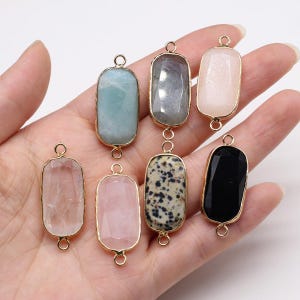 Puede incluir: Una colección de siete colgantes de piedras preciosas rectangulares, cada uno engastado en un marco de metal dorado con dos pequeños bucles circulares en la parte superior e inferior. Las piedras presentan varios colores, incluyendo azul claro, gris, rosa, negro y blanco moteado.