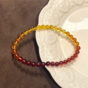 Peut inclure: Un bracelet de perles composé d'un dégradé lisse de perles sphériques translucides, passant d'un jaune miel vif à une extrémité à un rouge cerise profond et sombre à l'autre, assemblées sur un cordon flexible en une forme circulaire.
