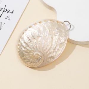 Op de afbeelding: Een ovaalvormige abalone schelphanger met een natuurlijke iriserende afwerking in wervelende crème, witte en parelmoerkleurige tinten. De schelp heeft een getextureerd oppervlak met een spiraalpatroon en een zilverkleurige metalen ring.