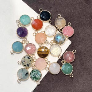 May include: A collection of round, faceted gemstone charms set in gold-toned metal frames. Each charm features a unique stone, including amethyst, tiger's eye, dalmatian jasper, and rose quartz, with two small loops on opposite sides for jewelry making.