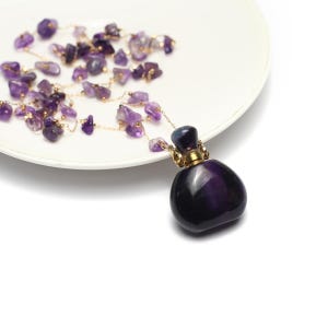 Peut inclure: Un flacon de parfum violet foncé avec un bouchon doré, relié à un collier de petites pierres d'améthyste. Le collier et le flacon reposent sur une assiette blanche. Le flacon a une finition brillante.