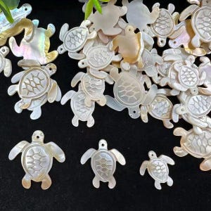 Puede incluir: Una colección de dijes de tortuga marina de nácar iridiscente, cada uno con un patrón de caparazón tallado y un pequeño orificio en la parte superior para su fijación. Los dijes muestran tonos naturales blanco nacarado y crema con acabado.