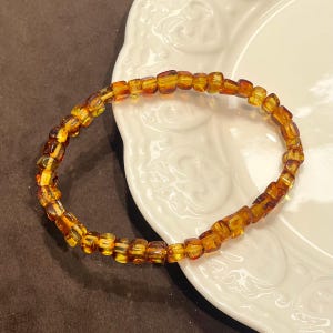 May include: A circular bracelet composed of small, polished, translucent amber beads in varying shades of golden yellow, orange, and deep brown. The beads are strung together in a continuous loop, showcasing natural variations in colour and clarity.