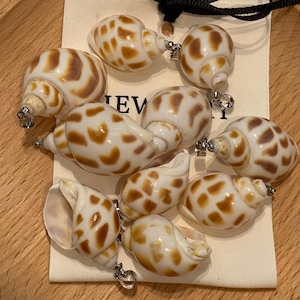 May include: A collection of nine white sea shell pendants featuring brown spotted patterns. Each shell is fitted with a silver-toned metal bail at the top for hanging. The shells are arranged on a cream-colored fabric tag with partial text visible.