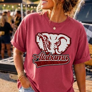May include: A crimson short-sleeve t-shirt featuring a graphic of a white elephant head with glittery details and the word Alabama in a script font. The shirt has a crew neckline and a relaxed fit, paired with light blue denim shorts.
