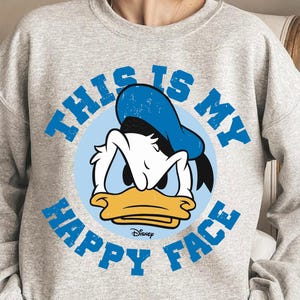 Peut inclure: Un sweat-shirt gris clair avec un dessin de Donald Duck et le texte "THIS IS MY HAPPY FACE" en bleu. Le logo Disney est en bas. Le dessin est centré sur le devant du sweat-shirt à col rond et à manches longues.
