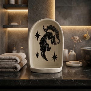 May include: Cream-colored ceramic soap dish with a black illustration of a stylized animal and stars. The dish has a curved, arched shape and is placed on a dark countertop in a bathroom setting.